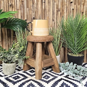 May include: A yellow ceramic mug with a white geometric pattern sits on a wooden stool. The stool has three legs and is made of natural wood. The mug is surrounded by green plants in pots.