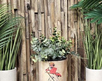 May include: A vintage-style metal planter with a Johnnie Walker label featuring a man in a red coat and white pants walking with a cane. The planter is filled with green plants and is sitting on a wooden surface.