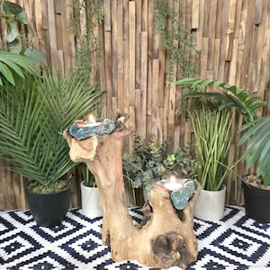 May include: A natural wooden candle holder with two teal glass candle holders, each with a lit white candle. The wood is light brown and has an organic shape. The background includes plants and a black and white patterned rug.