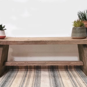 May include: A rustic wooden bench with a natural finish. The bench has a wide, flat top and two angled legs. It is sitting on a patterned rug. There are three potted plants on the bench, two with green leaves and one with a pink pot.