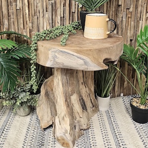 May include: A rustic wooden side table with a natural wood finish. The table has a round top and a thick, irregular base. A small potted plant and a mug with a geometric pattern are on the table.