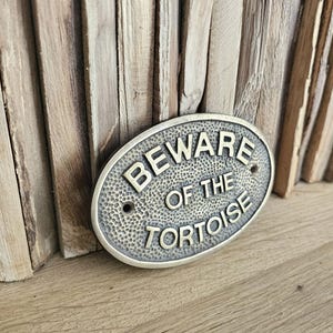 May include: Oval metal sign with the text "BEWARE OF THE TORTOISE" in capital letters. The sign has a textured surface and is mounted on a wooden background. The sign is a bronze colour.