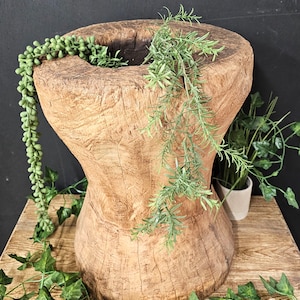 May include: A large, rustic wooden planter with a wide, round opening. The planter is filled with green plants, including a trailing succulent and a leafy sprig.