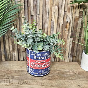 May include: A vintage-style metal can planter with a Coca-Cola design. The can is blue and red with white lettering and features the text "Delicious Refreshing Drink Coca-Cola 5¢ At All Soda Fountains". The planter is filled with a green leafy plant.