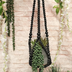 May include: Black wooden bead hanging planter with a green succulent plant inside. The planter is hanging from a brick wall.
