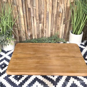 May include: A rectangular wooden cutting board with rounded edges. The board is a warm brown color and sits on a black and white patterned surface. Decorative plants in pots are in the background, with a wooden plank wall.