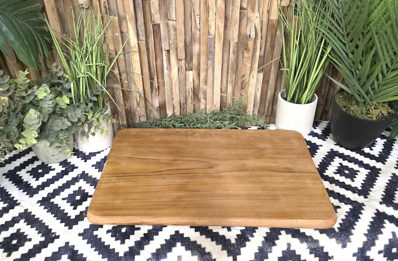 May include: A rectangular wooden cutting board with rounded edges. The board is a warm brown color and sits on a black and white patterned surface. Decorative plants in pots are in the background, with a wooden plank wall.