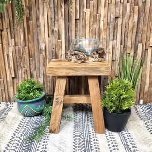 May include: A rustic wooden stool with a natural wood finish. The stool has a simple design with four legs and a rectangular seat. A clear glass bowl sits on top of a piece of driftwood on the stool. The stool is on a white and black patterned rug.