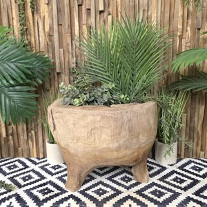 May include: A large, rustic wooden planter with three sturdy legs, filled with lush green plants. The planter has a natural, weathered appearance and is set against a backdrop of wooden planks and a black and white patterned rug.