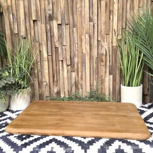 May include: A rectangular wooden cutting board with rounded edges sits on a black and white patterned surface. The background features a wooden wall and potted plants, creating a natural aesthetic.