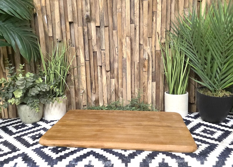 May include: A rectangular wooden cutting board with rounded edges sits on a black and white patterned surface. The background features a wooden wall and potted plants, creating a natural aesthetic.