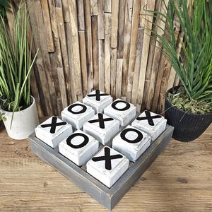 May include: A wooden tic-tac-toe game with white painted blocks and black X's and O's. The game is set on a grey wooden board.