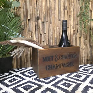 May include: A wooden champagne bottle holder with the words "MOET & CHANDON CHAMPAGNE" printed on the front. A black champagne bottle is in the holder. The holder is on a black and white patterned surface.