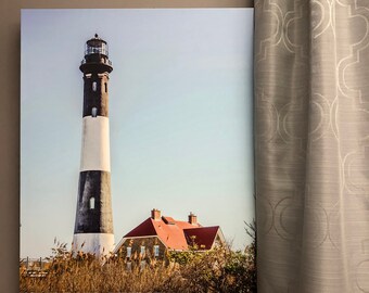 May include: A black and white lighthouse with a red roofed house in the foreground. The lighthouse is tall and slender with a white base and black stripes. The house is a small, one-story building with a red roof and white trim. The background is a blue sky with white clouds.
