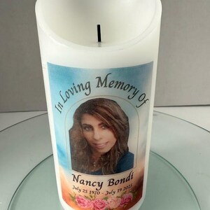 May include: White pillar candle with a printed label. The label reads "In Loving Memory Of" above a photo of a woman, with the name "Nancy Bondi" and dates "July 25 1970 - July 19 2025" below. Pink roses are at the bottom.