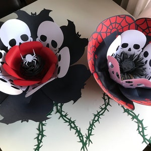 May include: Two paper flowers with a Halloween theme. One flower is black with red and white accents and features a spider in the center. The other flower is black with red and white spiderweb accents. Both flowers have a stem with thorns.