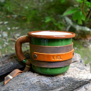 Op de afbeelding: Groene emaille mok met een bruin leren handvat en een bruine leren wikkel om de mok. De leren wikkel heeft het woord "KIEL" erop gegraveerd.