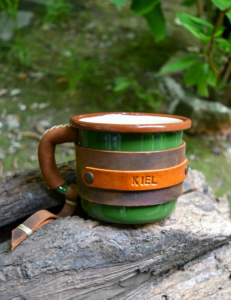 Op de afbeelding: Groene emaille mok met een bruin leren handvat en een bruine leren wikkel om de mok. De leren wikkel heeft het woord "KIEL" erop gegraveerd.