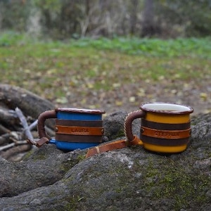 May include: Two personalized enamel mugs with leather handles. The mug on the left is blue with the name "Laura" on it. The mug on the right is yellow with the name "Stephanie" on it.