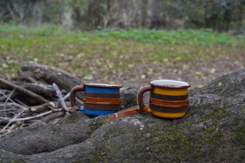 May include: Two personalized enamel mugs with leather handles. The mug on the left is blue with the name "Laura" on it. The mug on the right is yellow with the name "Stephanie" on it.