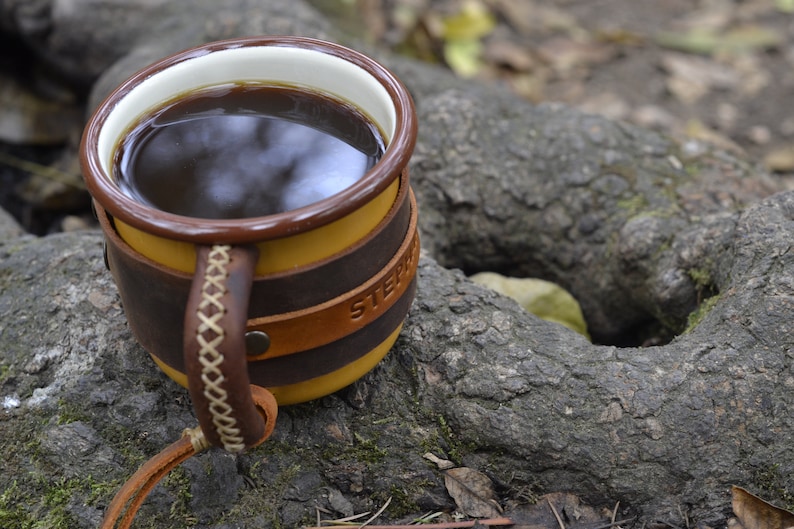 May include: A brown leather-wrapped mug with a yellow ceramic interior and a white rim. The mug is filled with dark coffee and has the word "STEPH" embossed on the side.