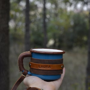 May include: A blue enamel mug with a brown leather handle and wrap. The leather wrap has the name "LAURA" embossed on it.