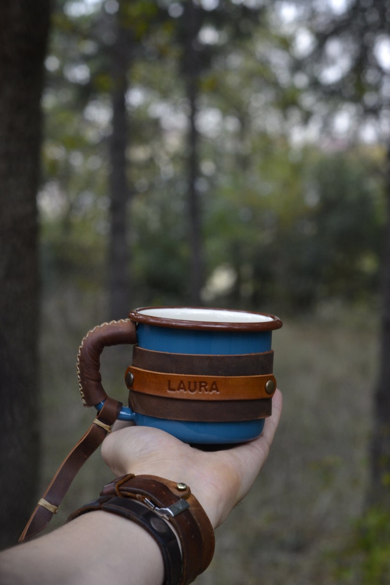 May include: A blue enamel mug with a brown leather handle and wrap. The leather wrap has the name "LAURA" embossed on it.