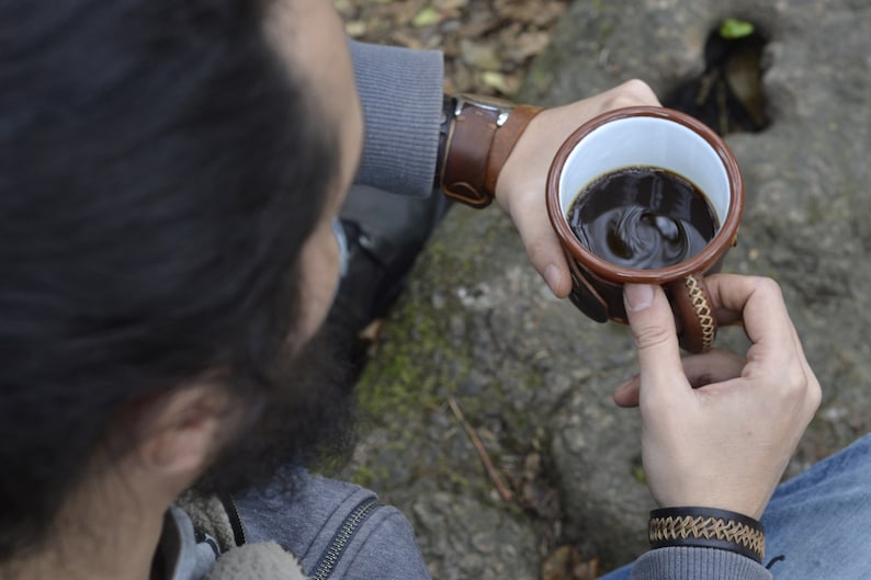 May include: A person holding a brown leather-wrapped mug with a white interior filled with coffee. The mug has a brown leather strap and stitching. The person is wearing a gray sweater and blue jeans.