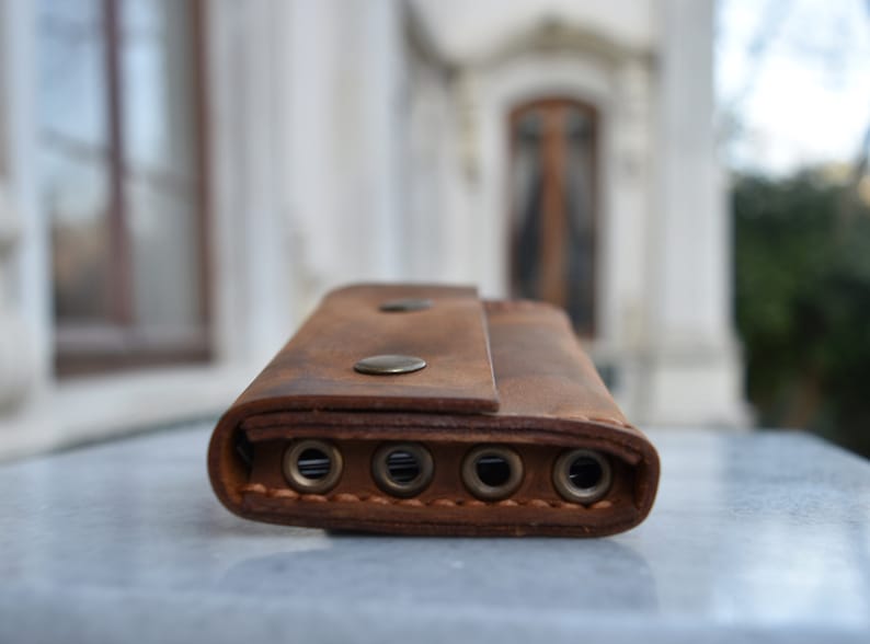 May include: A brown leather key holder with five key rings and two metal snaps. The key holder is on a white surface.