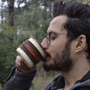 May include: A white ceramic mug with a brown leather wrap and the name "Kagan" embossed on it. The mug has a leather strap attached to the handle.