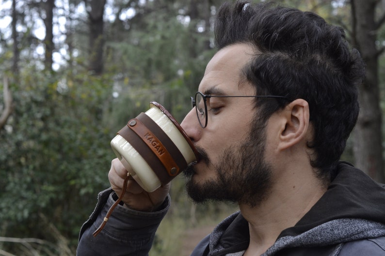May include: A white ceramic mug with a brown leather wrap and the name "Kagan" embossed on it. The mug has a leather strap attached to the handle.