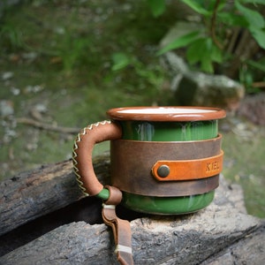 May include: Green enamel mug with a brown leather handle and a brown leather wrap around the mug. The leather wrap has a brown leather strap with a metal rivet and the word "KIEL" embossed on it.