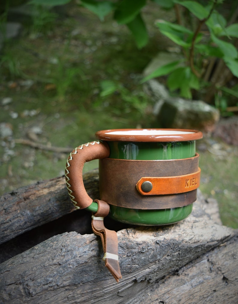 May include: Green enamel mug with a brown leather handle and a brown leather wrap around the mug. The leather wrap has a brown leather strap with a metal rivet and the word "KIEL" embossed on it.