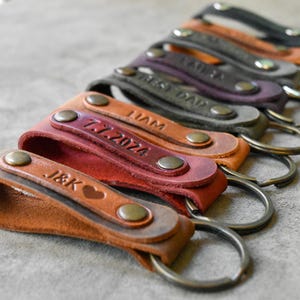 Puede incluir: Una colección de llaveros de cuero personalizados en varios colores, incluyendo marrón, rojo y gris. Cada llavero presenta un anillo metálico y está grabado con iniciales, fechas o nombres. Los llaveros están asegurados con remaches metálicos.