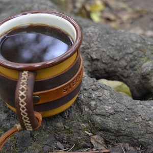 Op de afbeelding: Een bruine leren mok met een gele keramische binnenkant en een witte rand. De mok is gevuld met donkere koffie en heeft het woord "STEPH" in reli&euml;f aan de zijkant.