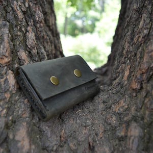 May include: A dark gray leather wallet with two brass snaps and a stitched edge. The wallet is lying on a tree trunk.
