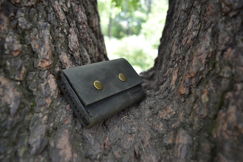 May include: A dark gray leather wallet with two brass snaps and a stitched edge. The wallet is lying on a tree trunk.