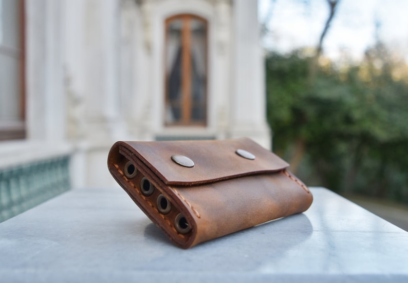 May include: A brown leather key holder with five key rings and two silver snaps. The key holder is lying on a white surface.