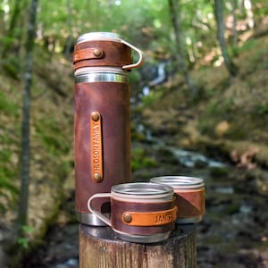 Op de afbeelding: Een bruine, met leer omwikkelde thermoskan met bijpassende deksel en twee kleine bekers. De thermoskan heeft de tekst "HUDSON FAMILY" op een bruine leren strip. De bekers hebben de tekst "JANIS". De items staan op een houten paal in een bos.