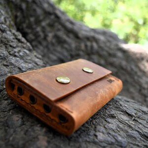 May include: A brown leather wallet with two brass snaps and multiple slots. The wallet is lying on a rough, textured surface.