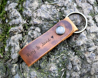 Könnte beinhalten: Ein brauner Leder-Schlüsselanhänger mit einem Metallring und einem silbernen Knopf. Der Schlüsselanhänger ist mit dem Text "Roy & Jane" und einem kleinen Herz graviert.