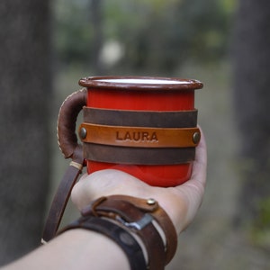 May include: Red enamel mug with a brown leather handle and wrap. The leather wrap has the name "LAURA" embossed on it.