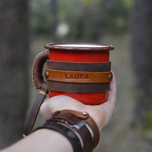 Op de afbeelding: Rode emaille mok met een bruin leren handvat en wikkel. De leren wikkel heeft de naam "LAURA" erop gegraveerd.