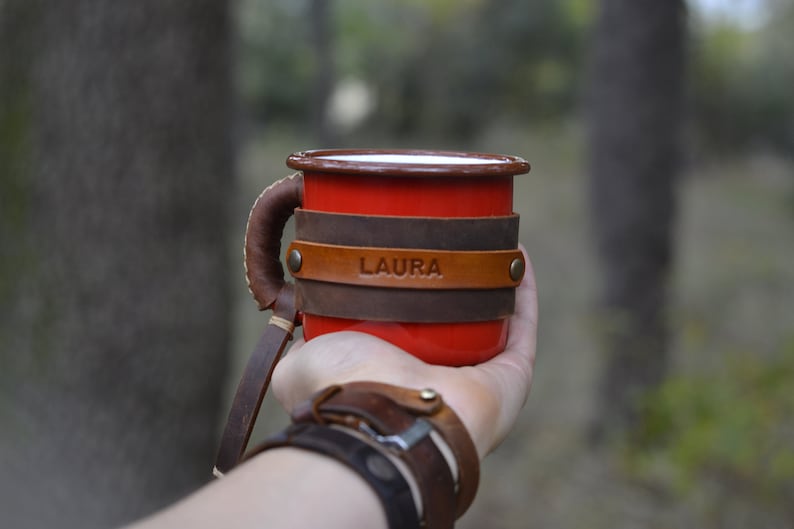 Op de afbeelding: Rode emaille mok met een bruin leren handvat en wikkel. De leren wikkel heeft de naam "LAURA" erop gegraveerd.