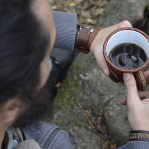 Op de afbeelding: Een persoon houdt een bruine leren mok vast met een witte binnenkant gevuld met koffie. De mok heeft een bruine leren band en stiksels. De persoon draagt een grijze trui en een blauwe spijkerbroek.