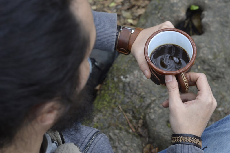 Op de afbeelding: Een persoon houdt een bruine leren mok vast met een witte binnenkant gevuld met koffie. De mok heeft een bruine leren band en stiksels. De persoon draagt een grijze trui en een blauwe spijkerbroek.