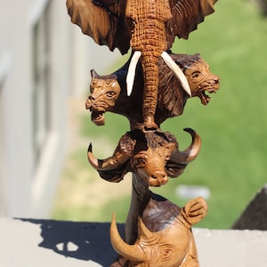 Può includere: Una scultura in legno di un elefante, un bufalo, un leone e un rinoceronte impilati uno sopra l'altro. La scultura è realizzata in legno scuro con accenti in legno chiaro.