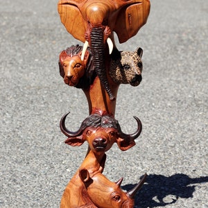 Op de afbeelding: Een houten sculptuur van vijf Afrikaanse dieren die op elkaar gestapeld zijn. De dieren zijn een olifant, een leeuw, een luipaard, een buffel en een neushoorn. De sculptuur is gemaakt van donkerbruin hout en heeft een natuurlijke houtfinish.