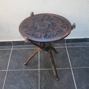 May include: A dark brown wooden folding table with a round top. The top is carved with a detailed design of animals and the word "Africa".