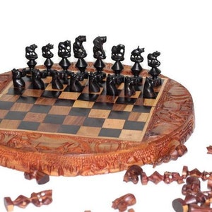 Può includere: Un set di scacchi in legno con pezzi neri e marroni. La scacchiera è rotonda e ha un design intagliato. I pezzi sono sparsi intorno alla scacchiera.